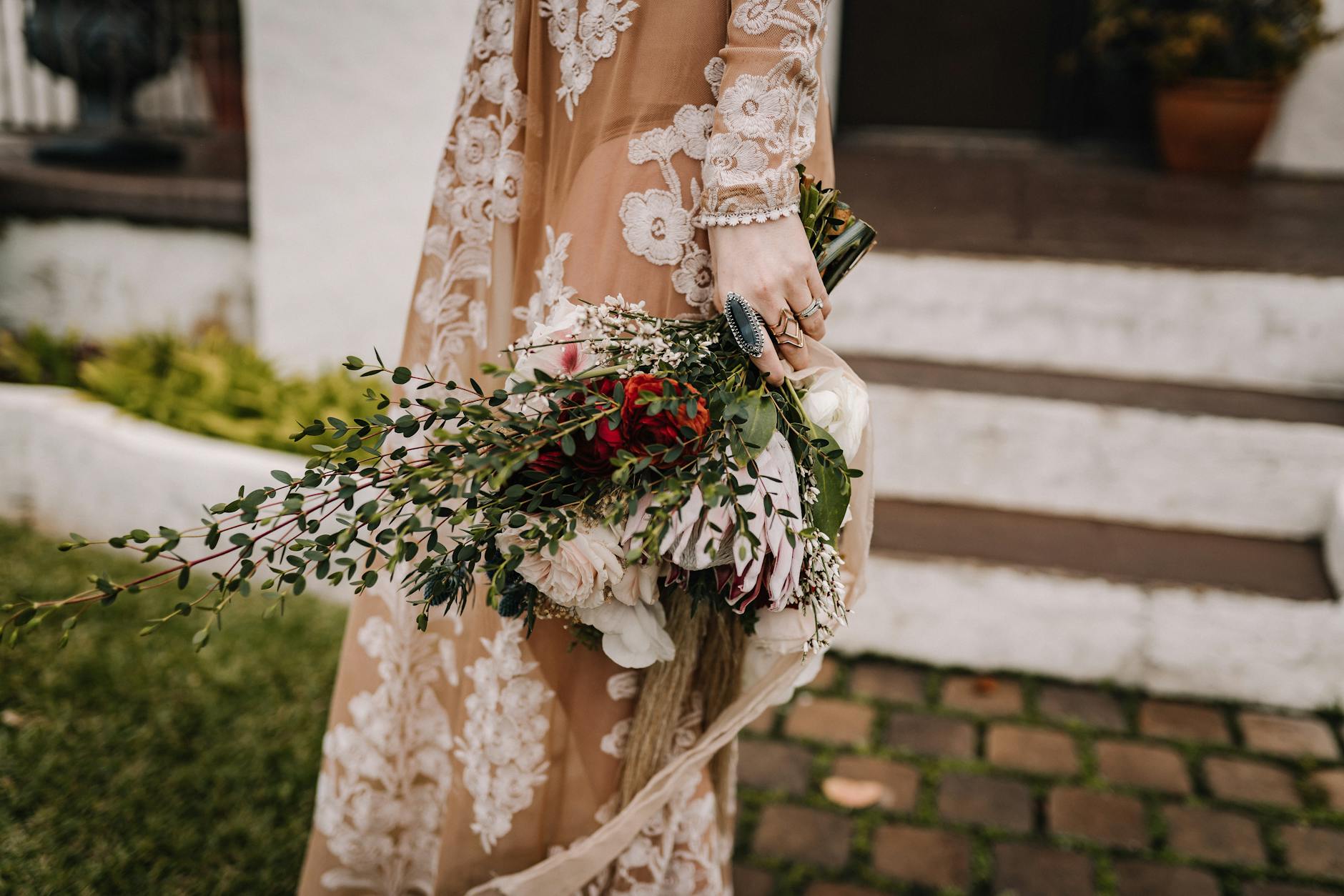 Biżuteria ślubna w stylu boho: co pasuje do koronki i kwiatów? Panna młoda z boho bukietem w dłoniach i koronkową suknią ślubną